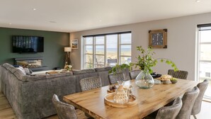 A modern living room with a large sofa, a wooden dining table, and a television. - 4 The Rocks - sleeps 8 in 4 bedrooms (Holywell Bay)