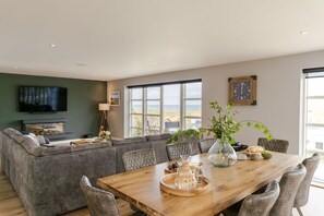 A modern living room with a large sofa, a wooden dining table, and a television. - 4 The Rocks - sleeps 8 in 4 bedrooms (Holywell Bay)