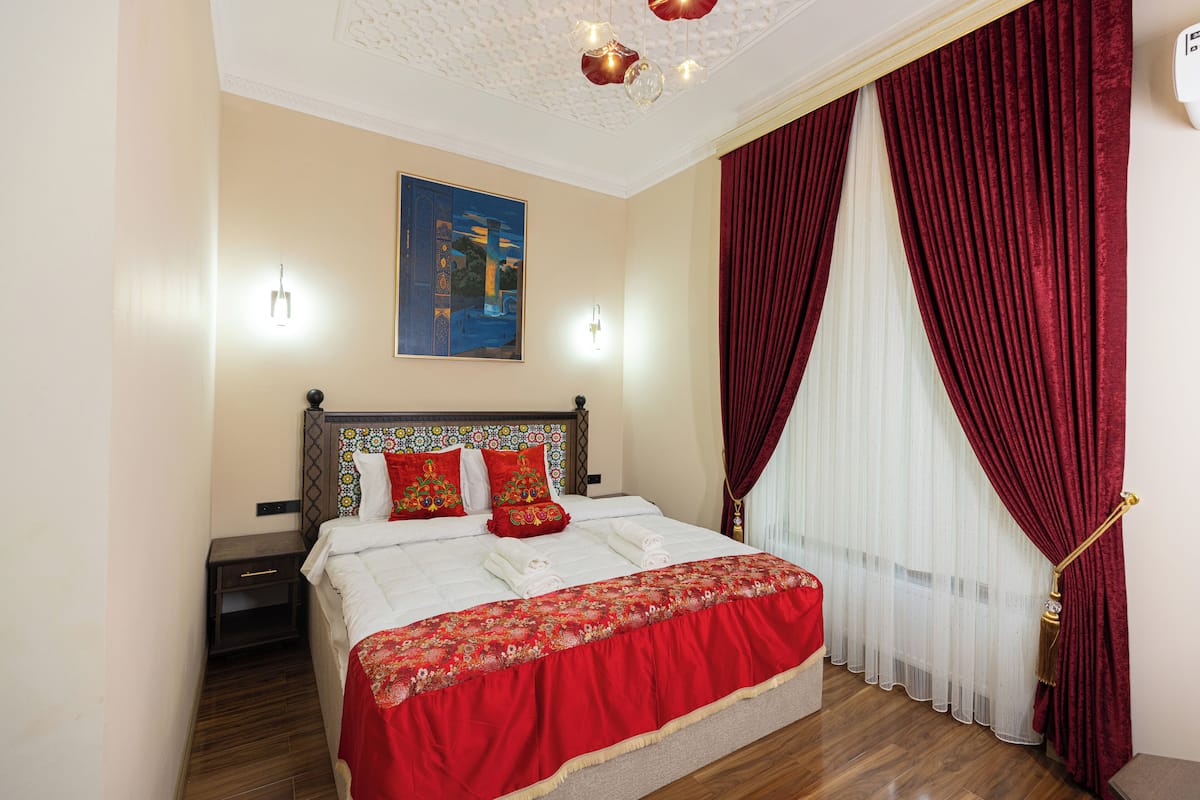 Une chambre d’hôtel avec un lit bien fait, une table de chevet, une tête de lit décorative, un tableau au mur et une fenêtre avec des rideaux rouges.