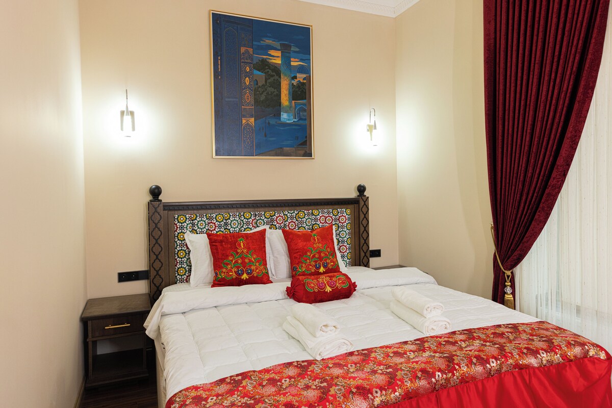 A neatly made bed with white linens and a red floral bedspread, a nightstand with a lamp, a wall-mounted painting, and a red curtain.