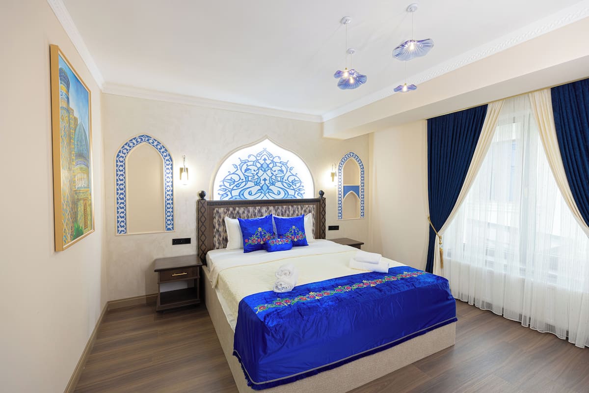 Une chambre d’hôtel avec un grand lit, une table de chevet, une commode, une arche décorative au mur et une fenêtre avec des rideaux.