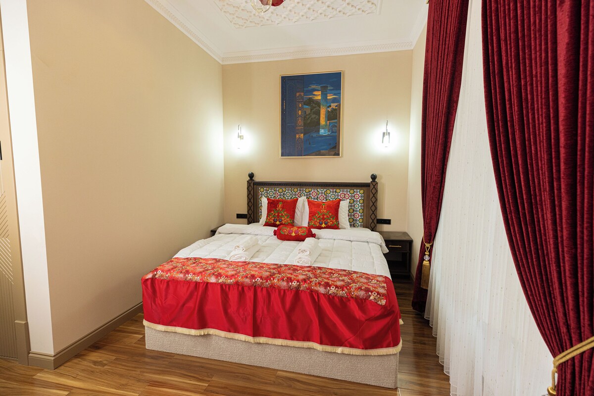 Une chambre d’hôtel avec un grand lit, une tête de lit ornée d’un motif floral, deux lampes de chevet, un tableau au mur et des rideaux rouges.