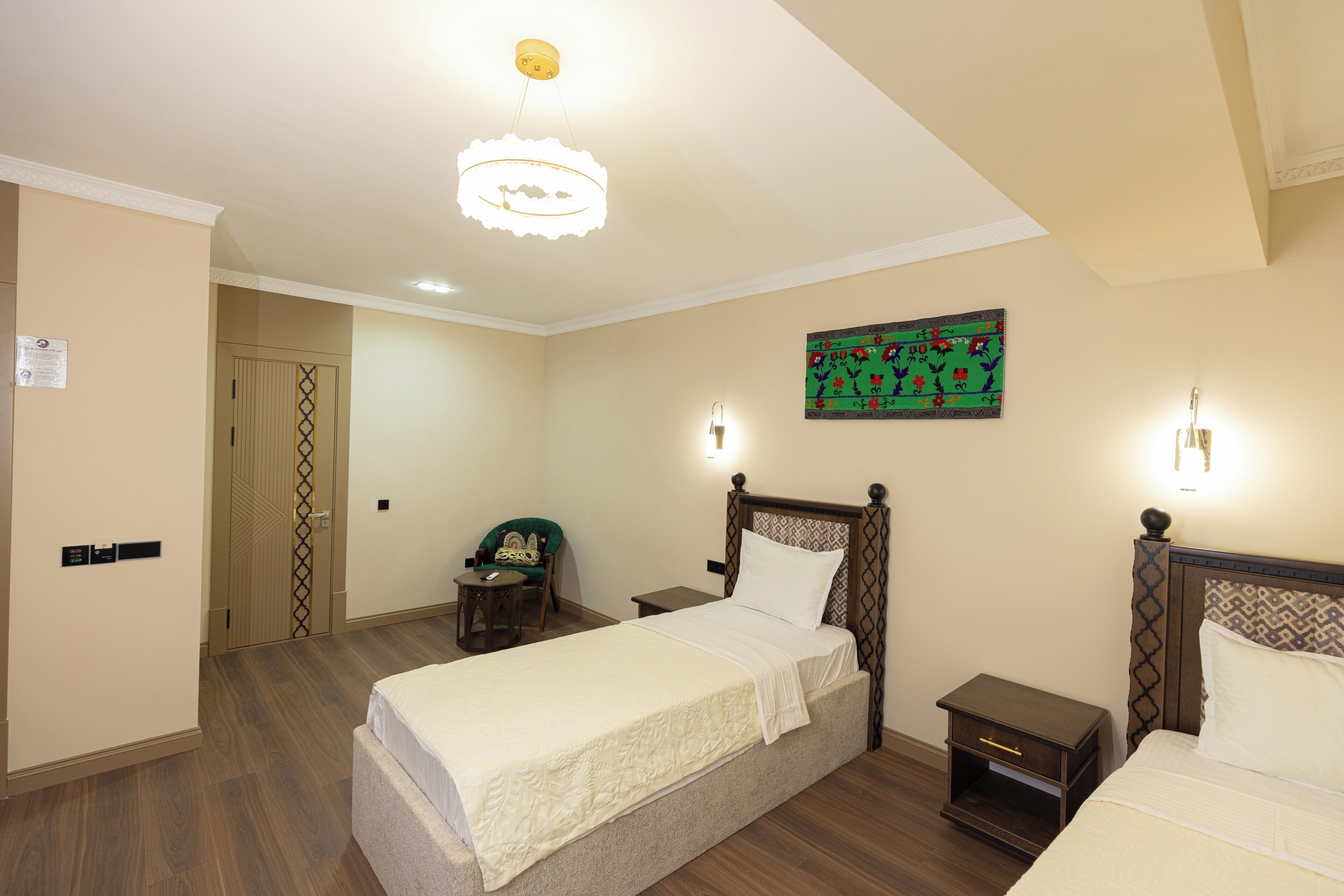 A hotel room with a bed, bedside tables, a chair, a painting on the wall, and a door leading to another room.