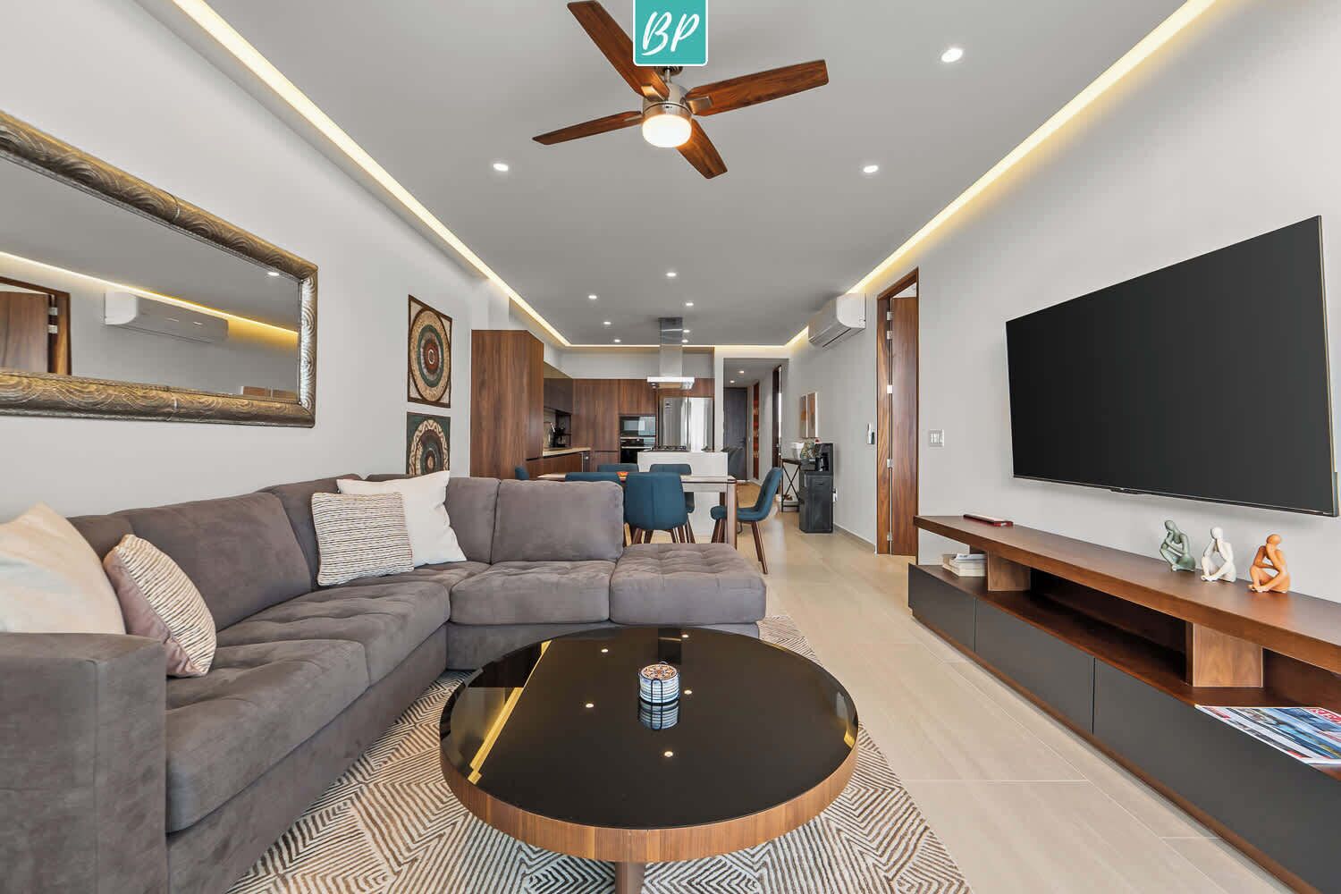 Un salon moderne avec un canapé d’angle, une table basse ronde, un téléviseur à écran plat et un ventilateur de plafond.
