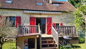 A stone building with a red door, a wooden deck, and a barbecue grill. - Charming gîte near Gourdon - stone, nature, enclosed garden, terrace (Concorès)