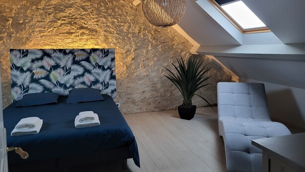 A bedroom with a sloped ceiling, a bed with a patterned headboard, a potted plant, a light-colored armchair, and a skylight. - Charming gîte near Gourdon - stone, nature, enclosed garden, terrace (Concorès)