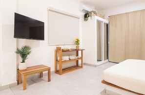 Une chambre d’hôtel avec un lit, un téléviseur fixé au mur, un petit banc en bois, une étagère avec des serviettes pliées, une plante en pot et une porte-fenêtre coulissante.
