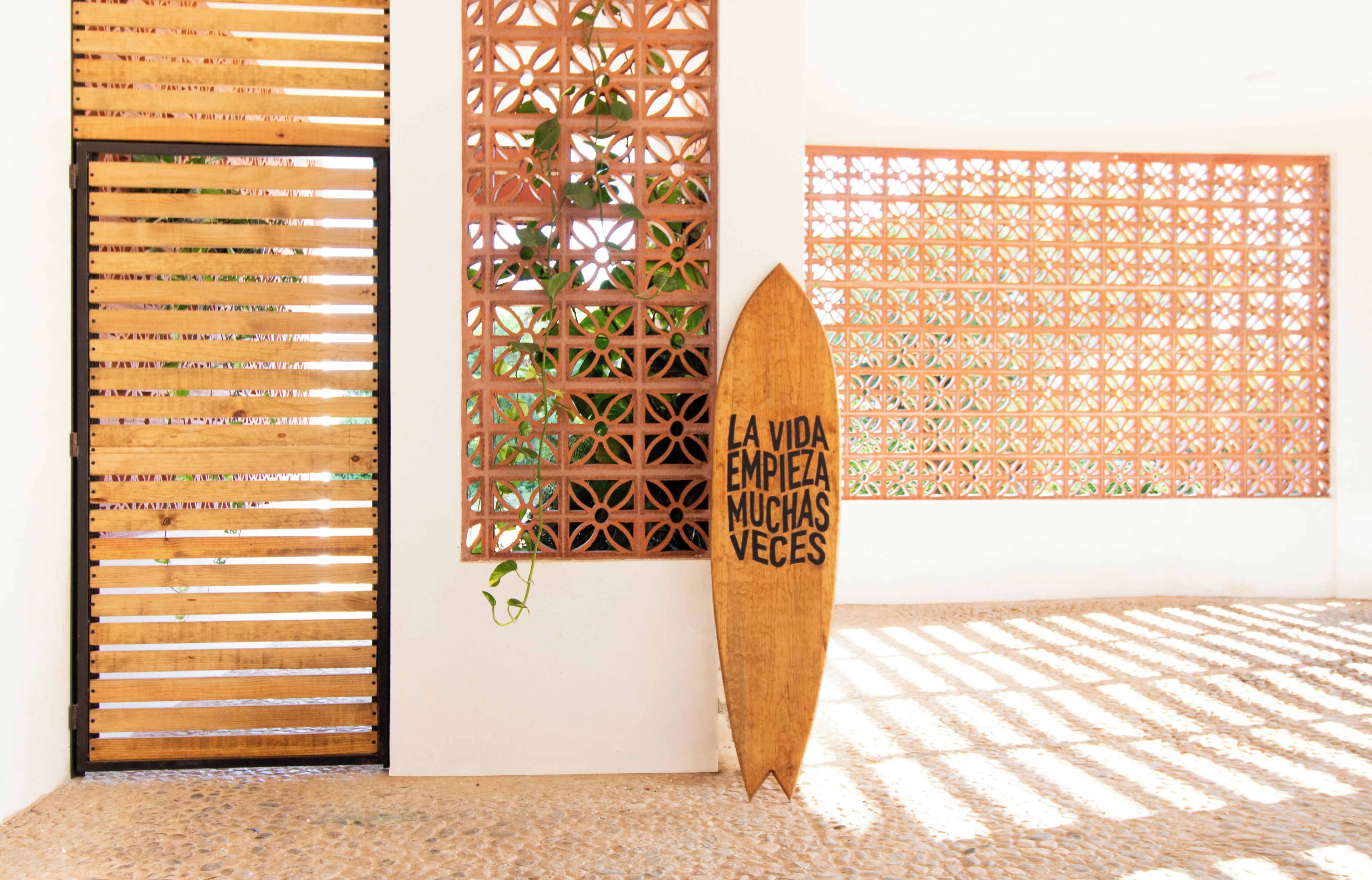 A wooden surfboard with text, a wooden slatted door, and a lattice wall with plants.