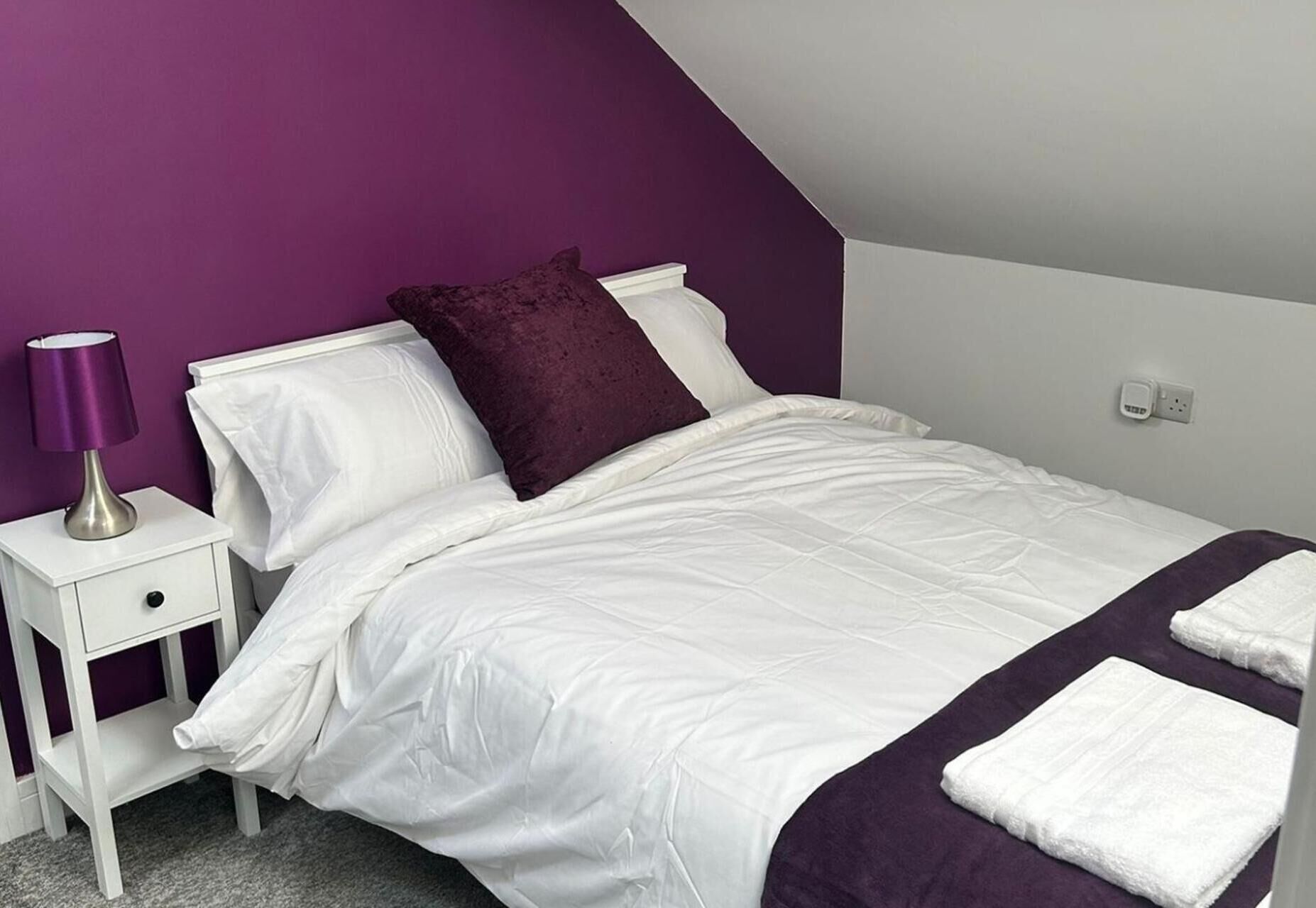 A bedroom with a bed, white nightstand, purple walls, and a white lamp.