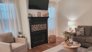 Een woonkamer met een open haard, een bank, een salontafel en een aan de muur bevestigde televisie.