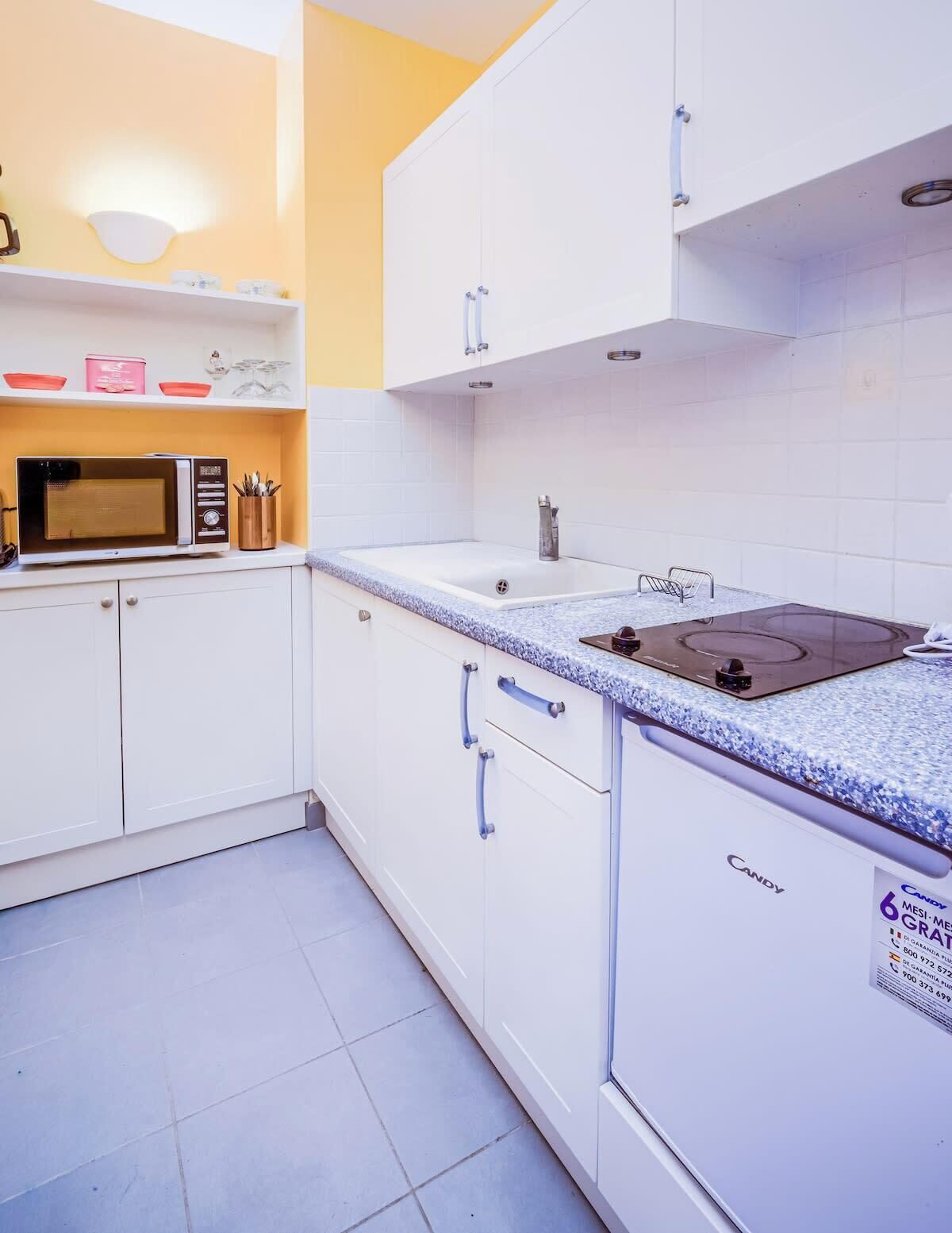 Een kleine keuken met witte kastjes, een magnetron, een spoelbak en een aanrecht met een waterkoker.