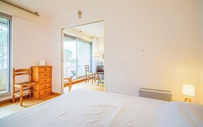 Ein Schlafzimmer mit einem Bett, einem Holzschrank, einem Stuhl, einem Heizkörper und einem Blick durch eine Tür auf einen Balkon.