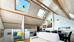 Ein Loft mit einer Couch, Esstisch und Stühlen, Holzbalken, Dachfenstern und einer Küchenzeile.