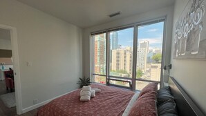 Een slaapkamer met een groot raam, een bed met een beddengoed met een patroon, en uitzicht op stadsgebouwen.