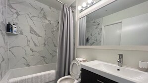 Een badkamer met een marmeren wand, een badkuip, een toilet, een wastafel en een spiegel met verlichting.