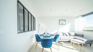 A modern living room with a round dining table, blue chairs, a grey sofa, and a large window. - La Vie en Bleu : T3 Lumineux (Guidel)