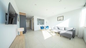A modern living room with a sofa, a TV, a dining area with chairs, and a staircase leading to another room. - La Vie en Bleu : T3 Lumineux (Guidel)