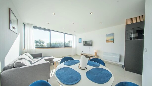 A modern living room with a sofa, round table, and chairs, a TV, and a kitchen area with a microwave. - La Vie en Bleu : T3 Lumineux (Guidel)