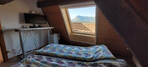 A small attic bedroom with a sloped ceiling, a bed with patterned bedding, a desk with a computer, and a window overlooking a mountainous landscape. - Panorama penthouse Du Lac (Sigriswil)