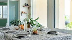 Mesa posta com pratos, copos e um pão, vista para a janela e plantas dentro de casa.
