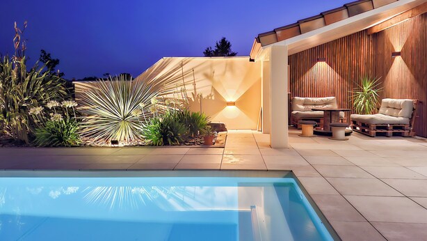 Piscina com área de terraço, incluindo uma mesa e cadeiras, cercada por plantas e um muro de madeira.