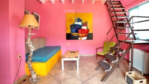 A pink room with a sofa, a small table, a lamp, a chair, a ladder, and a painting on the wall. - Oeve Apartment - A Colourful Loft in Kefalonia (Kefalonia)