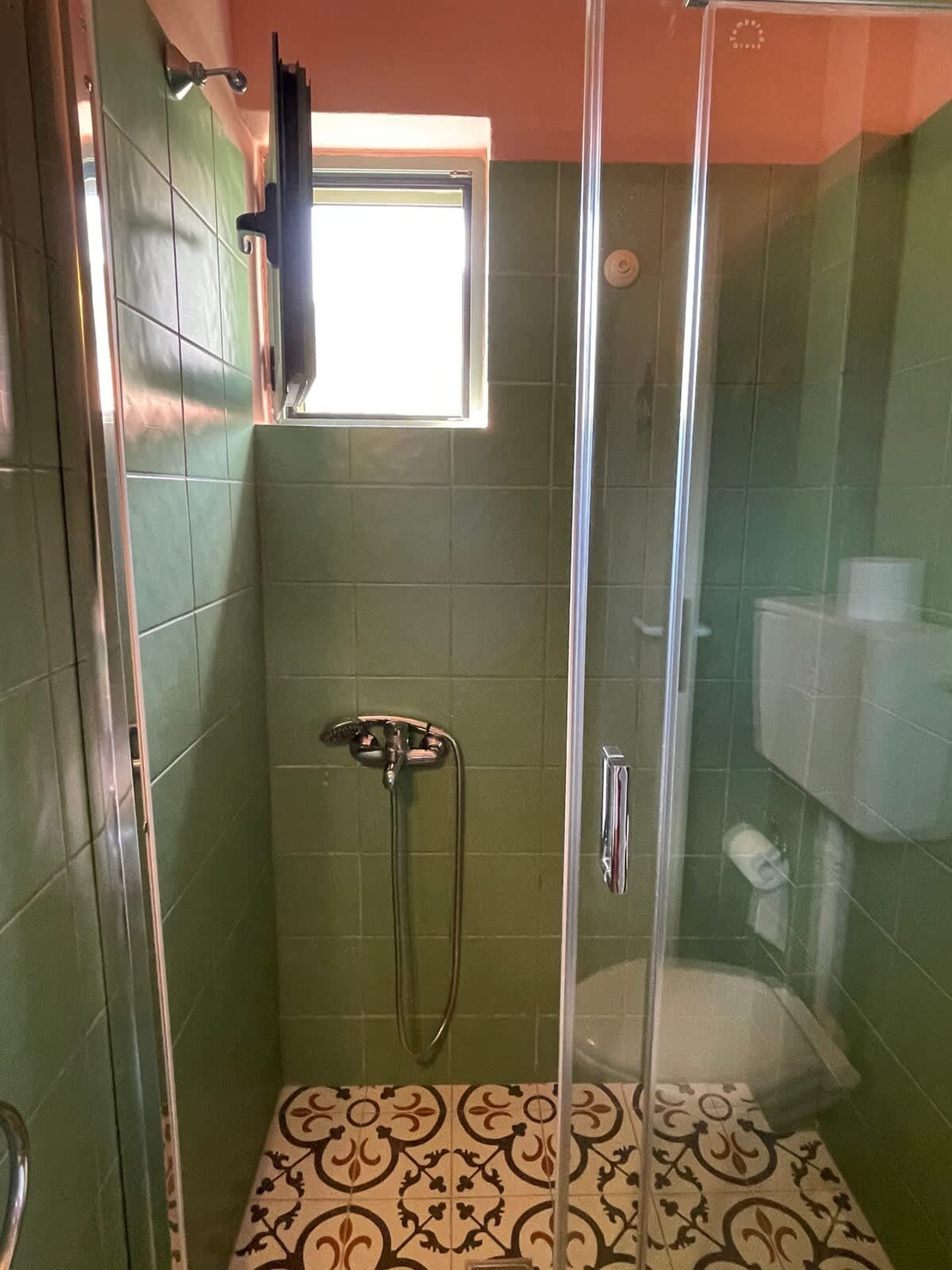 A glass-enclosed shower with a handheld showerhead, a small window, and patterned floor tiles.