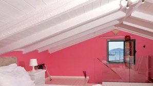 A bedroom with a sloped ceiling, a bed, a nightstand, a lamp, and a window with a view. - Oeve Apartment - A Colourful Loft in Kefalonia (Kefalonia)