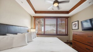 Ein Schlafzimmer mit einem Bett, einem Holzkopfende, einem Deckenventilator, einem Fenster mit Jalousien, einem Fernseher und einer Kommode.
