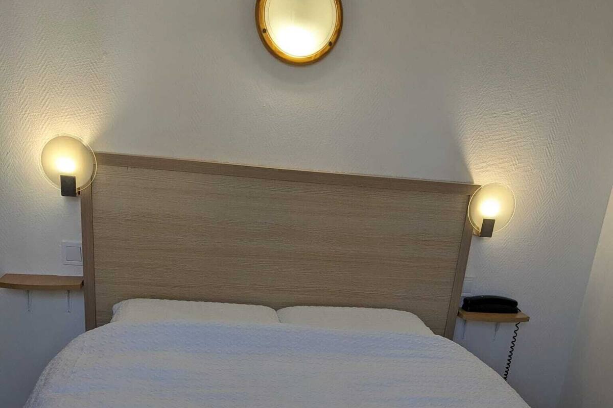 Sängyssä on puinen pääty, kaksi yöpöytävalaisinta ja pyöreä peili seinällä.