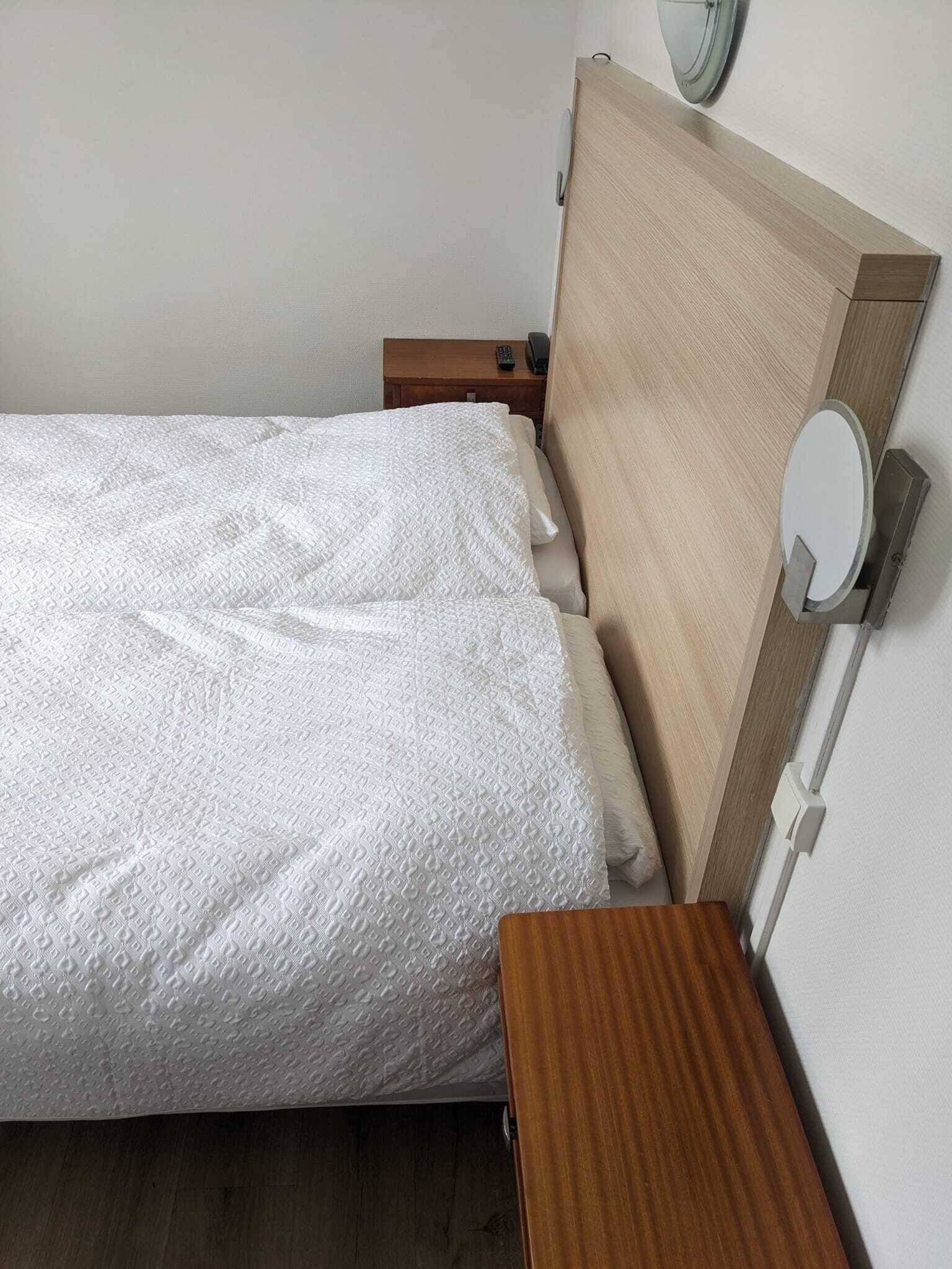 A neatly made bed with a white textured blanket, a wooden headboard, and a bedside table with a remote control.