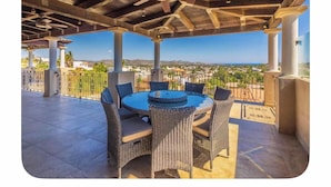 Une terrasse extérieure avec une table ronde et des chaises en osier, une vue sur un paysage côtier et une structure de plafond en bois.