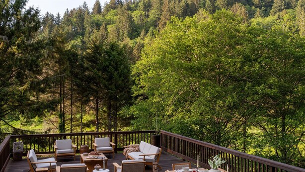 Une terrasse en bois, meublée pour l’extérieur, avec une table à manger pour quatre personnes et une vue sur une colline boisée.
