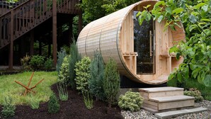 Une structure cylindrique en bois, dotée d’une porte, entourée d’un jardin où poussent diverses plantes, et à laquelle on accède par un escalier en bois menant à une terrasse.
