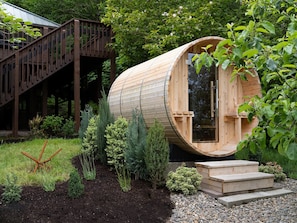 Ein zylindrisches Holzhäuschen mit Tür, umgeben von einem Garten mit verschiedenen Pflanzen und einer Holztreppe, die zu einer Terrasse führt.
