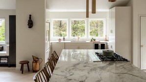 Une cuisine moderne dotée d’un îlot en marbre, de luminaires suspendus et offrant une vue sur les arbres grâce à de grandes fenêtres.