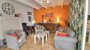 A living room with a sofa, chairs, a coffee table, a fireplace, a clock, and a kitchen area with a counter and cabinets. - Ker Monette - PORDIC (PORDIC)
