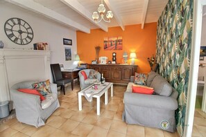 Ein Wohnzimmer mit einer Couch, Sesseln, einem Couchtisch, einem Kamin, einer Uhr und einer Küchenzeile mit Arbeitsfläche und Schränken.