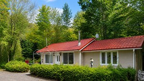 A single-story mobile home with a red roof, surrounded by dense green foliage and a gravel driveway. - Cozy Secluded Chalet with Hot Tub Surrounded by Nature (Val-Morin)