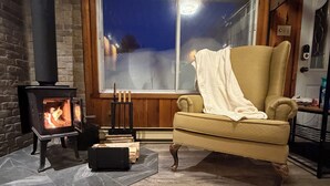 A cozy room with a stone fireplace, a comfortable armchair, and a large window. - Cozy Secluded Chalet with Hot Tub Surrounded by Nature (Val-Morin)