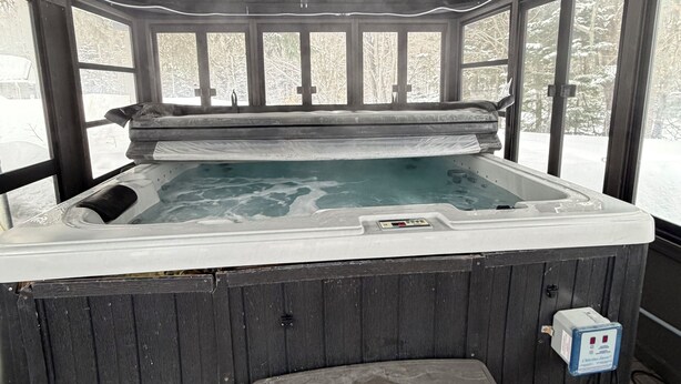 A hot tub with a wooden bench, a control panel, and a view of snow-covered trees through large windows. - Cozy Secluded Chalet with Hot Tub Surrounded by Nature (Val-Morin)