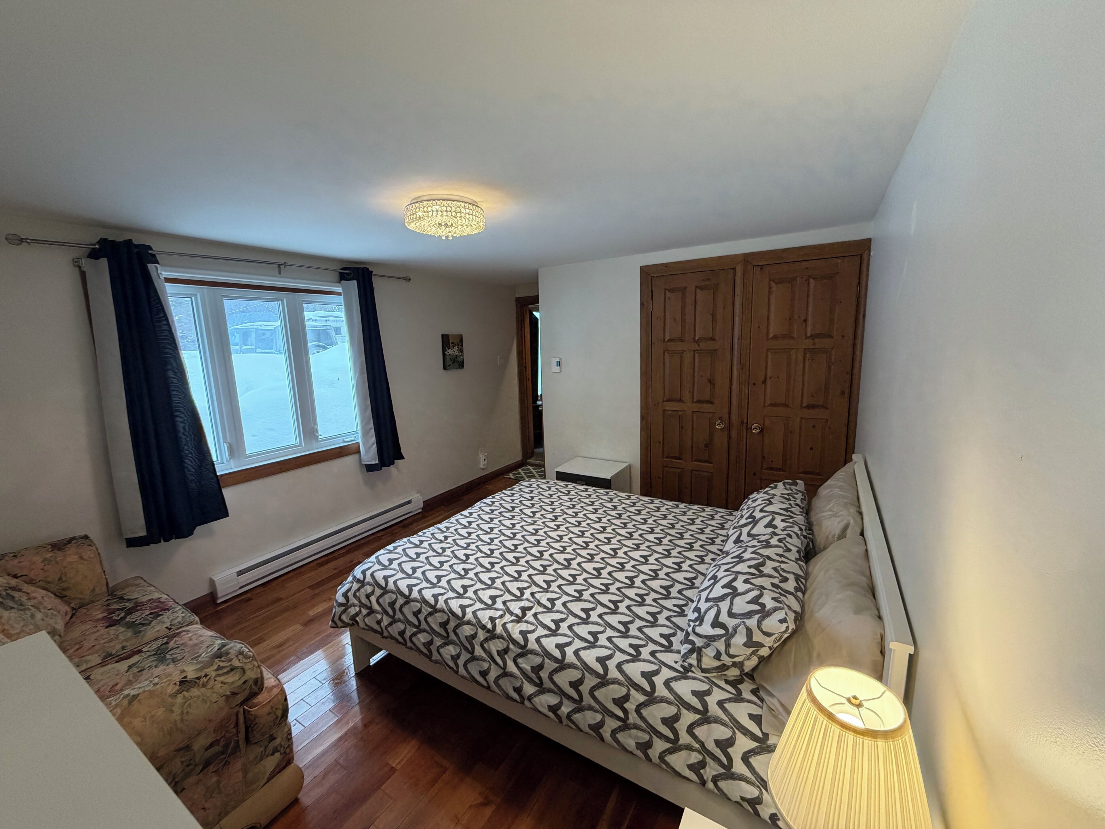 Une chambre à coucher comprenant un lit, une table de chevet, une lampe, une armoire, une fenêtre avec des rideaux et un radiateur.