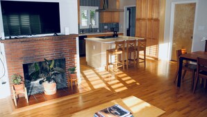 A living room with a fireplace, wooden flooring, a TV, and a kitchen area with wooden cabinets and a dining table.