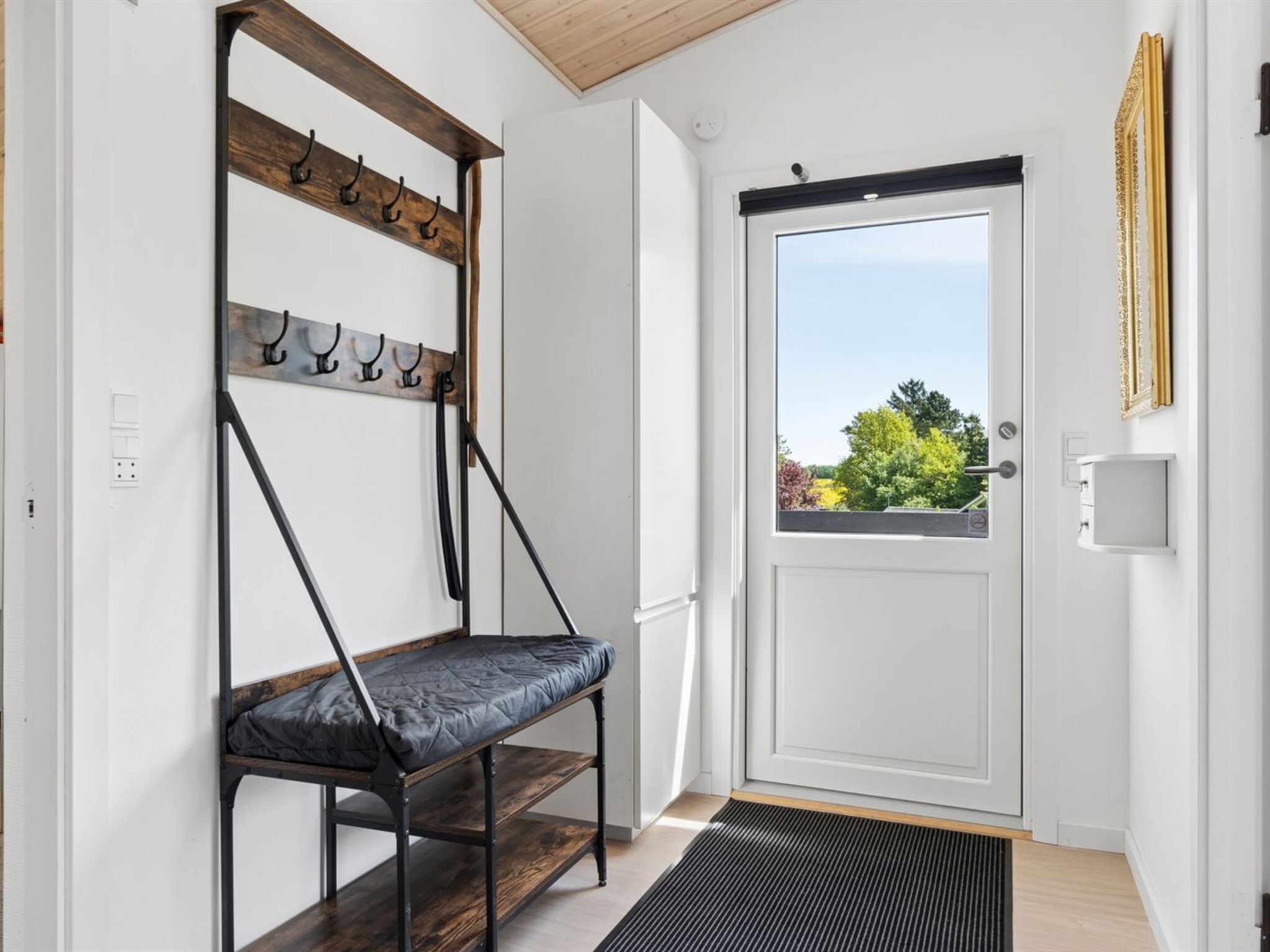 Una habitación con un banco, perchero, puerta con vistas y una estantería pequeña.