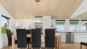 A modern kitchen with a wooden ceiling, a dining area with black chairs and a wooden table, and a view of greenery outside.