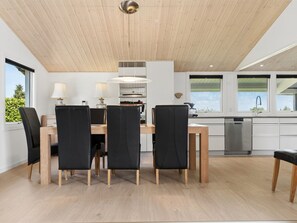 Eine moderne Küche mit Holzbalkendecke, einem Essbereich mit schwarzen Stühlen und einem Holztisch sowie Blick ins Grüne.