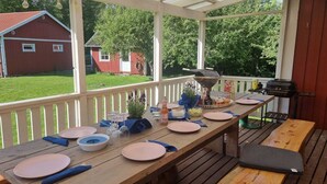A covered outdoor dining area with a table set for four, a grill, and a view of a red barn and green trees. - Family House Near Tiveden National Park with fireplace, piano, big property (Finnerödj)