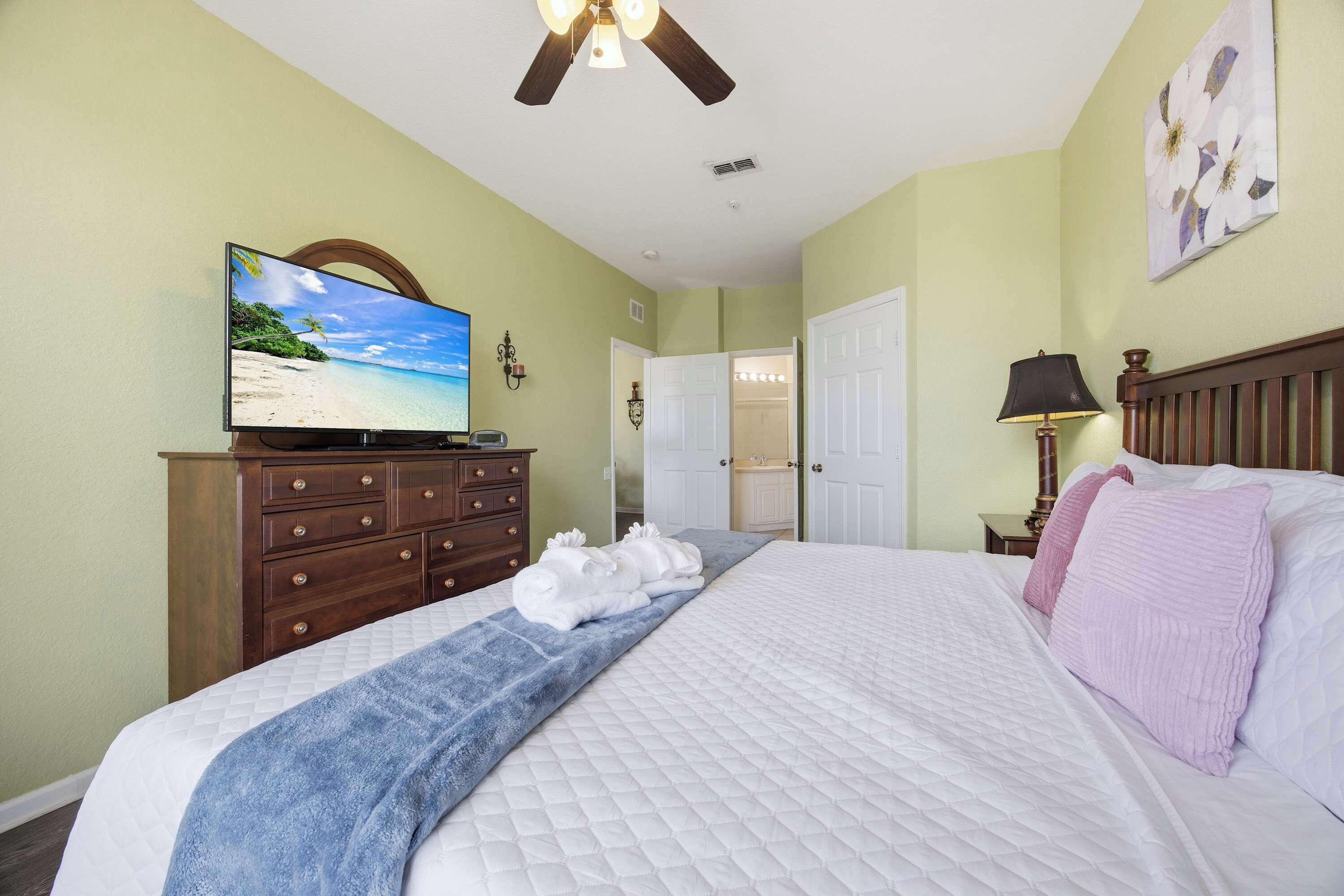 Un dormitorio con una cama, un tocador, un televisor, un ventilador de techo y una puerta que da a un baño.