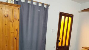 A bedroom with a bed, a wooden wardrobe, a grey curtain, and a door with vertical blinds. - Studio Apartment, Special Winter (Santa Teresita)