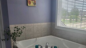 Une salle de bain avec une baignoire, une fenêtre avec des stores, une plante en pot et un tableau au mur.